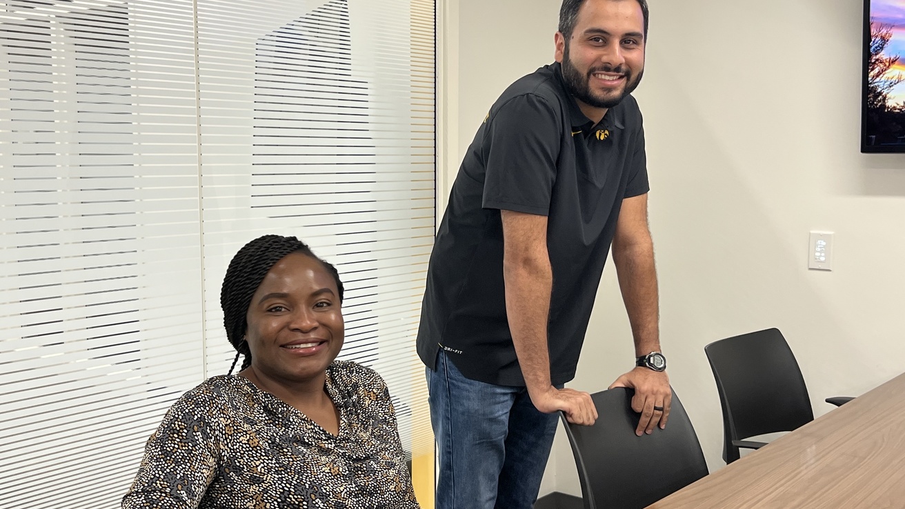 Two students - one is sitting, the other is leaning against a chair. They are both smiling. 
